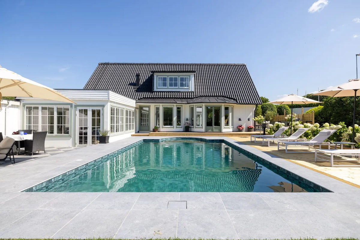 Pool framför en vit villa med stenlagd terrass, solstolar och parasoller